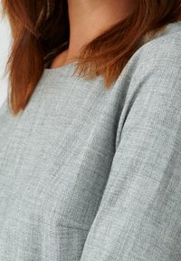 Donna con capelli rossi alla lunghezza delle spalle che indossa un maglione grigio chiaro con trama, mostrata dalla nuca fino a metà torso.