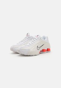 Fehér Nike sportcipők sima felsőrésszel, perforált részletekkel, fekete pipa logóval és piros díszítéssel a saroknál és a talpnál.