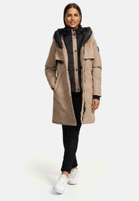 Parka longue beige matelassée avec une couche interne noire, dotée d'une capuche, de boutons sur le devant, de poches latérales et d'une fermeture éclair. Des baskets blanches complètent le look.