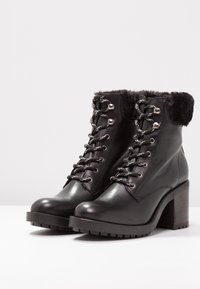 Botas de tobillo de cuero negro con un puño de piel sintética, parte delantera con cordones, accesorios plateados y tacón alto y grueso. Suela exterior con dibujo grueso para un mejor agarre.
