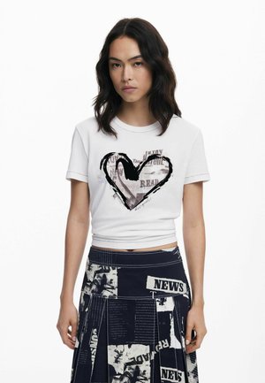 White cropped t-shirt with a black heart graphic and text, paired with a dark printed skirt featuring newsprint patterns and pleats.