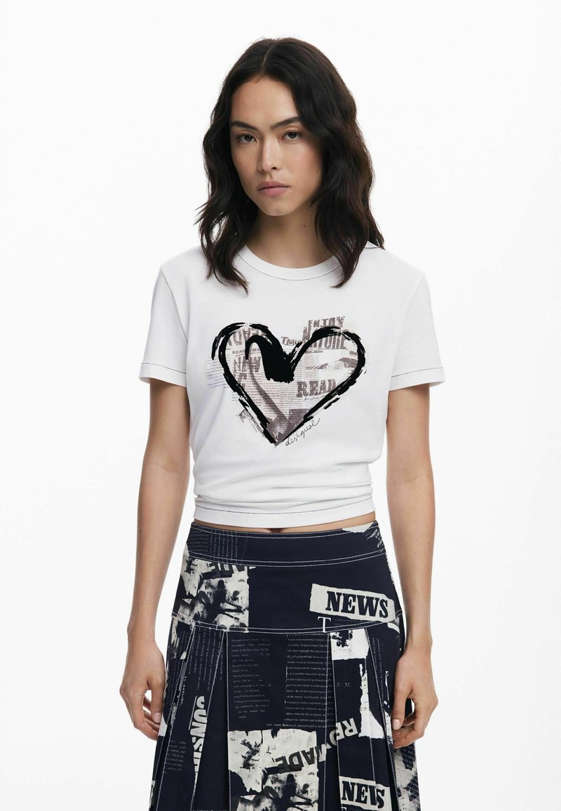 Weißes, kurzes T-Shirt mit einem schwarzen Herz-Graphic und Text, kombiniert mit einem dunklen, bedruckten Rock mit Zeitungsmuster und Falten.