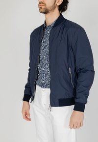 Giacca bomber navy in tessuto leggero con polsini e colletto a costine, dotata di una zip anteriore e tasche laterali. Camicia bianca a fantasia sotto.