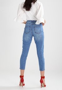 Džínové kalhoty s vysokým pasem v modré barvě, s tapered nohavicemi, pěti kapsami a viditelným prošitím, doplněné červenými sandály na vysokém podpatku.