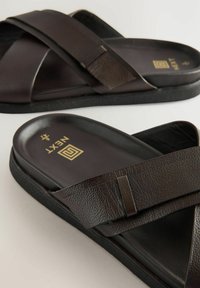 Sandales en cuir marron avec des brides croisées, une finition texturée et une semelle en caoutchouc. Détail du logo doré sur la semelle intérieure. Design plat.