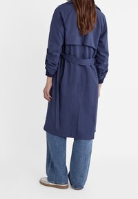 Marineblauwe trenchcoat met lange mouwen, een ceintuur om de taille en dubbele achterflappen; gestyled met blauwe jeans met wijde pijpen en casual schoeisel.