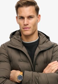 Mørkebrun puffjakke med hætte og lynlås, lagd over en struktureret sort sweater. Et guld- og blå ur bæres på håndleddet.