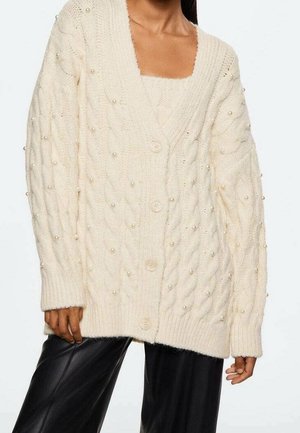 Femme portant un cardigan écru oversize en maille torsadée avec des ornements en perles et de gros boutons, associé à un pantalon en cuir noir.