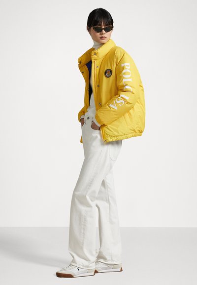 Geel pufferjack met een logo patch, witte letters op de mouw, gecombineerd met witte spijkerbroeken en sneakers. Model draagt donkere zonnebril.