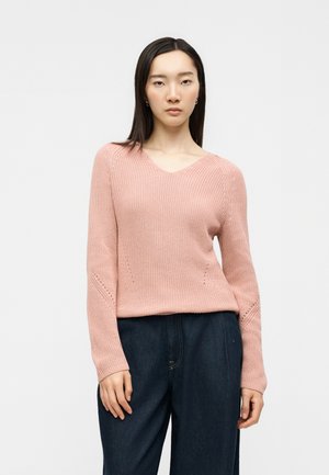 Mladá žena s rovnými tmavými vlasmi nosí svetloružový pletený sveter s výstrihom do V a tmavomodré nohavice s vysokým pásom, stojí na bielom pozadí.