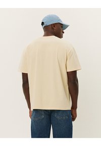 Tricou din bumbac bej cu o croială lejeră, mâneci scurte și guler rotund. Asortat cu blugi din denim albastri, văzut din spate.