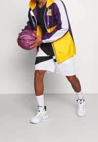 Lilla og gul vindjakke, sort skjorte, hvide shorts med sorte detaljer, holder en lyserød basketball med et mønster. Hvide sneakers.