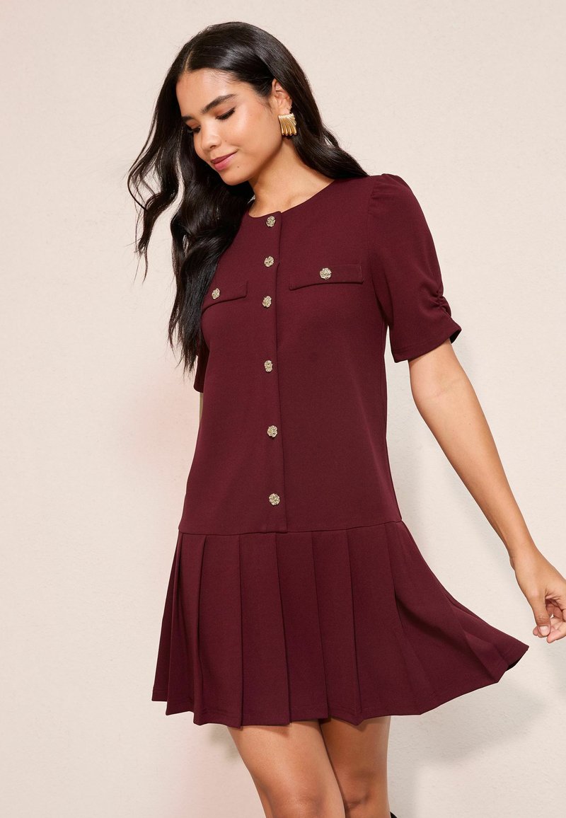 Rochie scurtă burgundy cu mâneci scurte și fustă plisată, cu decolteu rotund și accente cu nasturi aurii. Două buzunare frontale. Material neted.