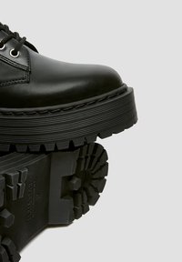Botte noire en cuir à lacets avec semelle épaisse et rainurée, vue de côté et de dessous sur fond gris uni.