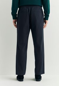 Donkerblauwe broek met elastische tailleband, twee achterzakken en een rechte pijpvorm. De stof lijkt glad en licht van gewicht. Schoeisel is donker van kleur.