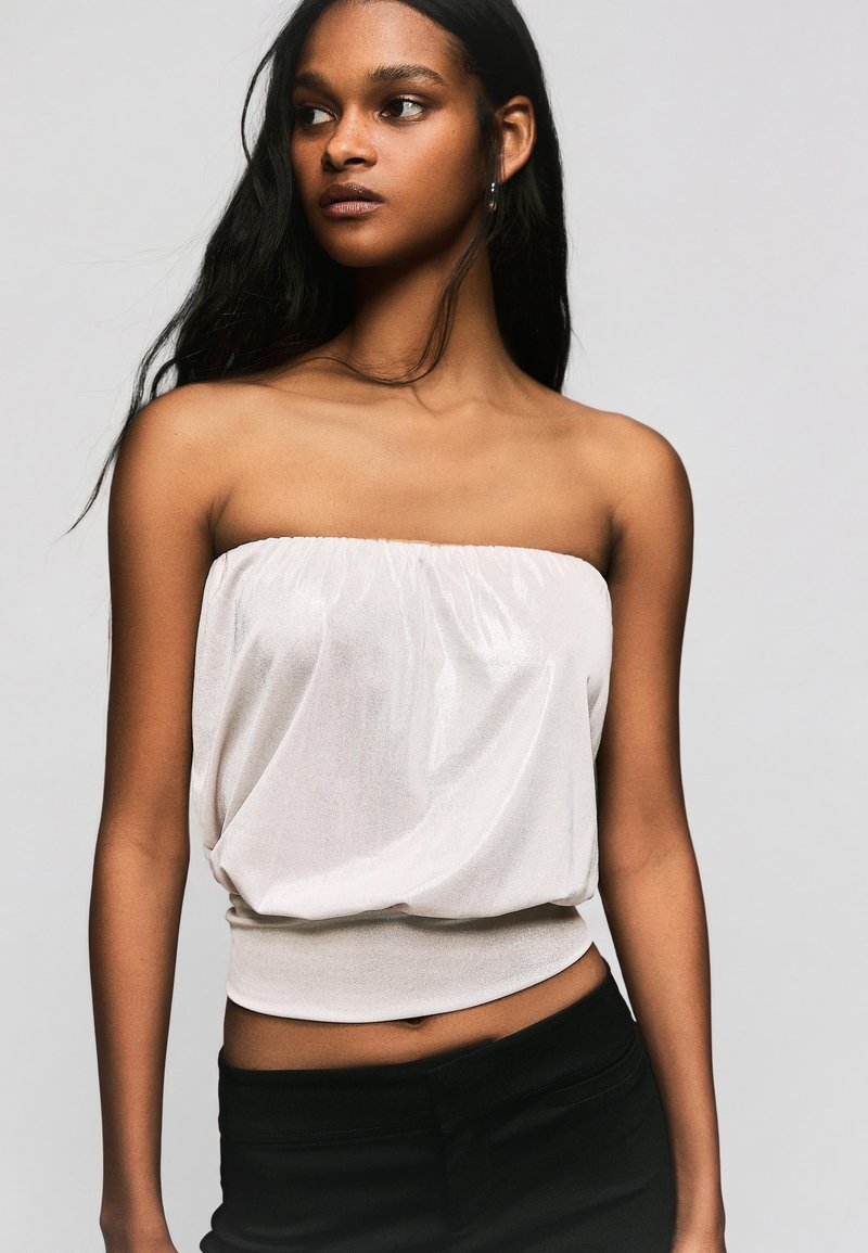 Femme aux longs cheveux noirs portant un haut blanc sans bretelles et un pantalon noir, regardant sur le côté sur un fond gris uni.