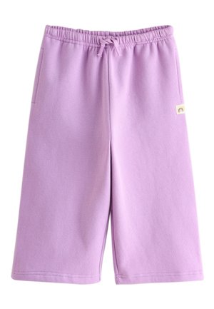 Lille barn står ved siden af en beige sofa iført en lavendelfarvet lynlåssweatshirt, matchende bukser og hvide sneakers.