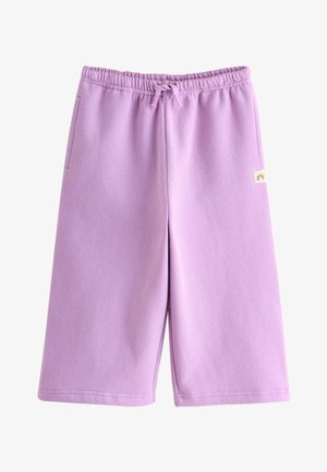 Lille barn står ved siden af en beige sofa iført en lavendelfarvet lynlåssweatshirt, matchende bukser og hvide sneakers.