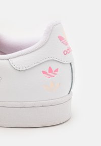 White sneaker with a smooth leather texture, featuring pink and light orange Adidas logo accents near the heel, and a textured rubber sole.