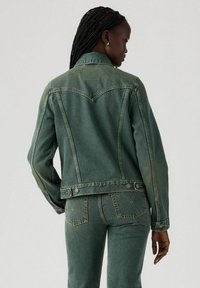 Green denim jacket with a classic design, featuring a pointed collar, back yoke seam, and buttoned cuffs. Textured fabric with visible stitching.