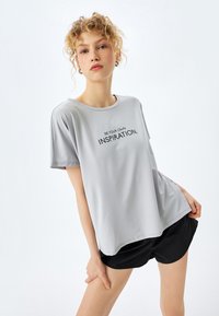 Camiseta de manga corta gris claro hecha de tejido suave que presenta el texto "SÉ TU PROPIA INSPIRACIÓN". Pañalones cortos deportivos negros completan el conjunto.