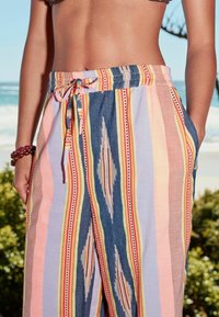 Dámský trup oblečený v barevných pruhovaných kalhotách s vázáním v pase a hnědém bikinovém topu, stojící venku poblíž pláže s oceánem a zelení.