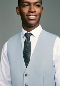 Gilet bleu clair porté sur une chemise blanche, associé à une cravate à motifs foncés ornée de fleurs bleues, mettant en valeur un tissu texturé et des doubles boutons.