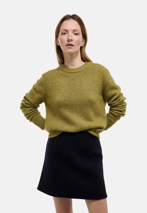 Pull vert en maille avec un col rond et des poignets côtelés, associé à une jupe noire trapèze. Le pull présente une finition texturée.