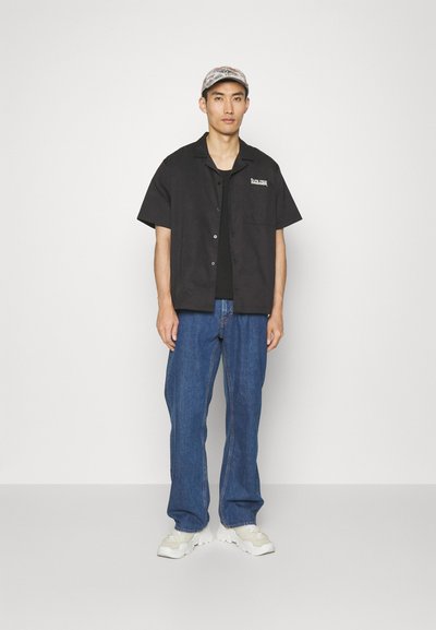 Melnas īsām piedurknēm pogājamas krekls, zem tā melna t-krekls ar apaļu kakla izgriezumu, zila džinsu bikses un baltas kedas. Cepure ar rakstu uz malām.