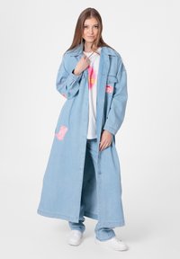 Veste en denim surdimensionnée bleu clair avec des patchs roses, portée sur un t-shirt graphique blanc et un pantalon en denim assorti. Des baskets blanches complètent le look.