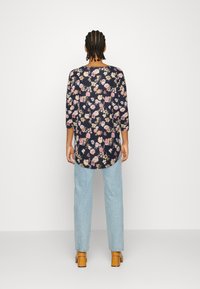 Blommig tunika med lång ärm i marin bakgrund, prydd med rosa och gula blommor, tillsammans med ljusblå jeans och beige klackskor.