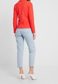 Czerwona dopasowana kurtka z kołnierzem, w połączeniu z jasnoniebieskimi dżinsami. Modelka ma na sobie białe szpilki, które prezentują minimalistyczny design.