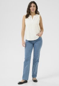 Blouse blanche sans manches avec des pois noirs, décolleté en V, associée à un jean droit bleu clair et des ballerines noires.
