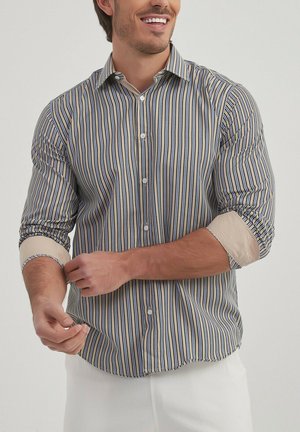 Chemise à manches longues avec des rayures verticales bleu, jaune et blanc. Présente un devant à boutons et des poignets beige retroussés. Tissu léger.