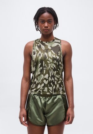 Žena stojaca čelom vpred, oblečená v zelenej maskáčovej bezrukávovej blúzke a lesklých olivovo zelených športových šortkách na jednoduchom pozadí.