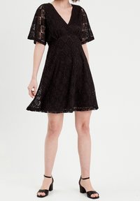 Robe en dentelle noire avec un décolleté en V et des manches courtes, présentant un motif floral, une jupe évasée et un tissu texturé. Sandales à talons blocs en daim.