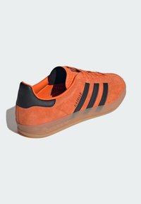 Adidași din suede portocalie cu dungi și accente negre, având o talpă din cauciuc și o căptușeală interioară portocalie. Brandingul "Gazelle" este vizibil pe lateral.