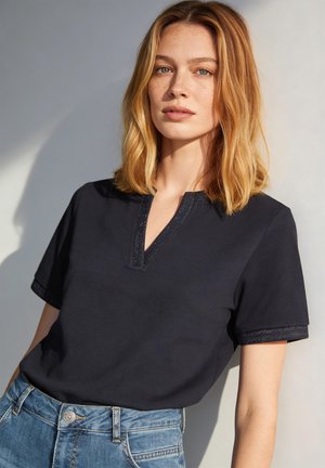 Jeune femme aux cheveux blonds mi-longs portant un haut noir à manches courtes et un jean bleu, debout devant un fond clair.