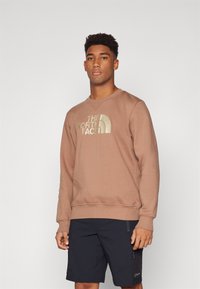 Felpa marrone in cotone con collo tondo e polsini a coste, caratterizzata da un grande logo dorato sul davanti. Indossata con pantaloni corti scuri.