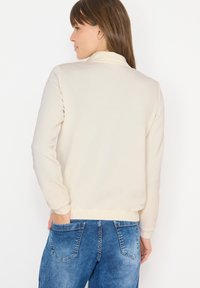 Sudadera color crema con cuello alto, puños y dobladillo acanalados. Llevada con jeans de mezclilla azul que presentan bolsillos traseros visibles.