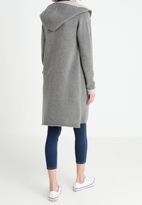 Grå huvtröja med en mjuk textur, långa ärmar och en medellång längd. Bärs över blå jeans och vita sneakers, vilket visar en avslappnad stil.