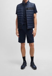 Gilet matelassé marine avec des coutures verticales, écusson logo noir ; associé à un short marine assorti et des baskets, créant une tenue coordonnée.