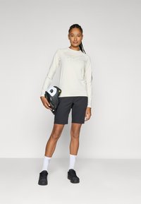 Chemise longue beige clair avec logo, shorts noirs, et un casque noir avec des accents blancs tenu à la main. Chaussures noires et chaussettes blanches présentées.
