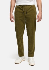 Uomo che indossa pantaloni in corduroy verde oliva con vita regolabile e sneakers bianche, in piedi contro uno sfondo neutro.