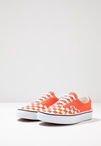 Orange lærredssneakers med et hvidt ternet mønster på tåen og siderne, prydet med hvide snørebånd og en sort gummisål.