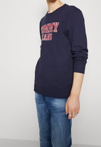 Navy sweatshirt with "TOMMY JEANS" in red lettering. Features ribbed cuffs and hem, with a relaxed fit paired with light denim jeans.