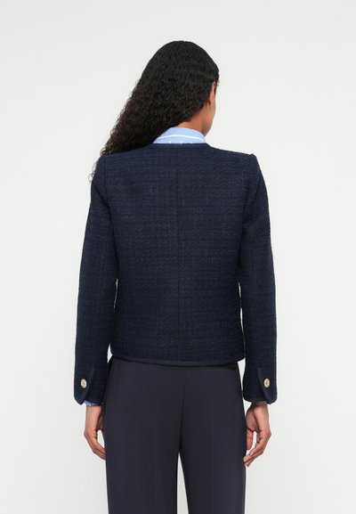 Giacca di tweed blu navy con design strutturato, scollatura rotonda, maniche lunghe e polsini abbottonati, abbinata a pantaloni scuri.