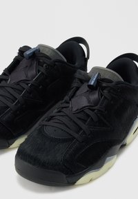 Par de zapatillas negras de corte bajo con parte superior texturizada, cordones ajustables con tope, collar acolchado y detalles en la suela blanca sobre un fondo claro.