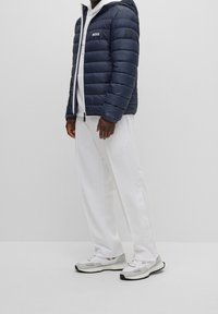 Muž v námořnické prošívané bundě, bílé mikině s kapucí, bílých kalhotách s postranními knoflíky a bílých teniskách na jednotném světlém pozadí.