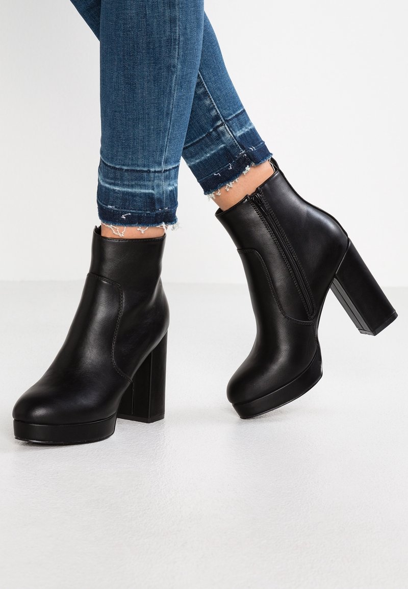 Bottines compensées en cuir noir avec talons épais portées avec un jean skinny bleu à ourlet effiloché sur fond clair.